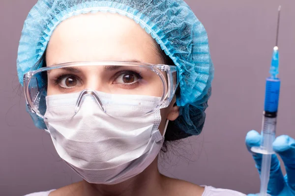 beautiful young girl in medical mask wearing glasses and gloves is holding a syringe with blue liquid