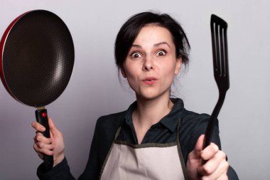 Önlüklü duygusal kadın yemek hazırlıyor, bulaşıkların elinde, gri arka planda.