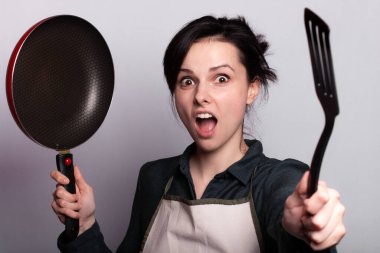 Önlüklü duygusal kadın yemek hazırlıyor, bulaşıkların elinde, gri arka planda.
