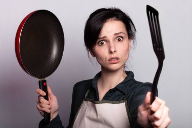 Önlüklü duygusal kadın yemek hazırlıyor, bulaşıkların elinde, gri arka planda.