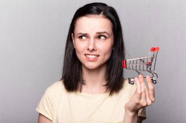 Elinde küçük bir market arabası tutan güzel bir kadın, gri bir arka plan.