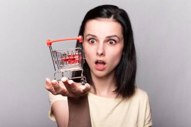 Elinde küçük bir market arabası tutan güzel bir kadın, gri bir arka plan.