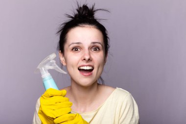 Koruyucu eldivenli güzel bir kadın gri arka planda bir deterjan ve toz bezi tutuyor.