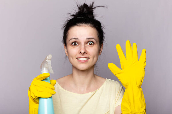 Beautiful young woman in protective gloves is holding a detergent and a duster, on gray background