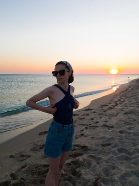 Güneş gözlüklü kadın gün batımında sahilde duruyor.