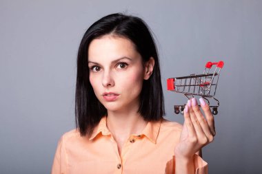 Turuncu gömlekli kadın elinde küçük bir alışveriş arabası tutuyor.