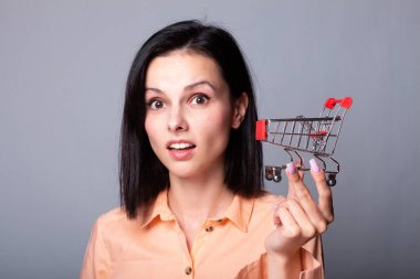 Turuncu gömlekli kadın elinde küçük bir alışveriş arabası tutuyor.