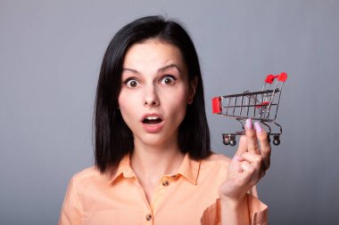 Turuncu gömlekli kadın elinde küçük bir alışveriş arabası tutuyor.