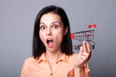 Turuncu gömlekli kadın elinde küçük bir alışveriş arabası tutuyor.