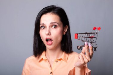 Turuncu gömlekli kadın elinde küçük bir alışveriş arabası tutuyor.