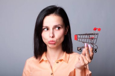 Turuncu gömlekli kadın elinde küçük bir alışveriş arabası tutuyor.