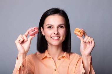 Turuncu tişörtlü gülümseyen kadın elinde Fransız kurabiyeleri, gri arka plan.