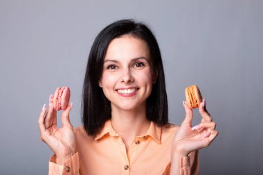 Turuncu tişörtlü gülümseyen kadın elinde Fransız kurabiyeleri, gri arka plan.