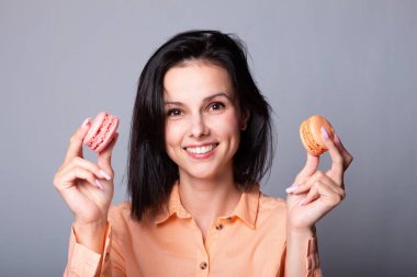 Turuncu tişörtlü gülümseyen kadın elinde Fransız kurabiyeleri, gri arka plan.