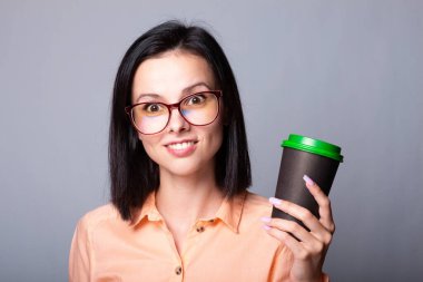 Turuncu tişörtlü ve elinde bir fincan kahve tutan gri arka planlı bir kız öğrenci.