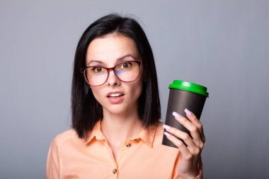 Turuncu tişörtlü ve elinde bir fincan kahve tutan gri arka planlı bir kız öğrenci.