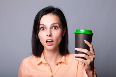 Turuncu gömlekli tatlı kız elinde bir fincan kahve tutuyor, arka planı gri.