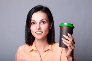 Turuncu gömlekli tatlı kız elinde bir fincan kahve tutuyor, arka planı gri.