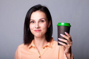 Turuncu gömlekli hoş bayan ellerinde bir fincan kahve tutuyor, gri arka planda.