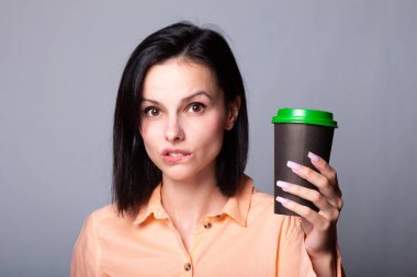 Turuncu gömlekli hoş bayan ellerinde bir fincan kahve tutuyor, gri arka planda.