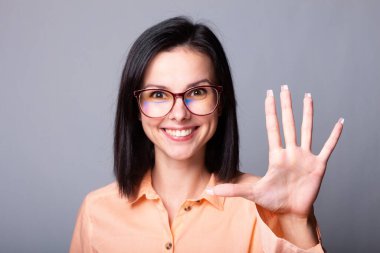 Turuncu tişörtlü kadın beş parmak gösteriyor, gri arkaplan.