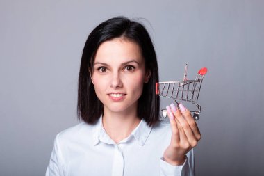 Beyaz gömlekli gülümseyen kadın elinde küçük bir alışveriş arabası, finans sembolü.