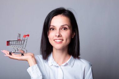 Beyaz gömlekli kadın elinde küçük bir alışveriş arabası tutuyor.