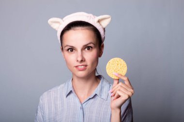 Taze derili ve genç görünümlü, gözü kozmetik süngerle kaplanmış, gri günlük gömlek giyen, güzellikle ilgilenen, beyaz arka planda izole edilmiş genç bir kadın.