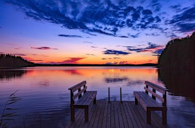 Two wooden bench or chairs on a wood dock facing a lake at sunset in Finland clipart