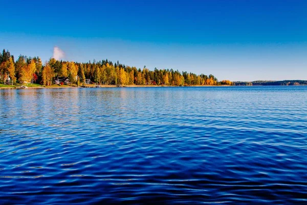 Podzimní listí, barevný Les nad modrým jezerem s červenými chatami ve Finsku. — Stock fotografie