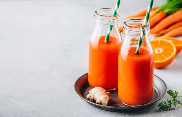 Свежая морковь, имбирь и апельсиновый напиток в стеклянных бутылках на сером каменном фоне — стоковое фото