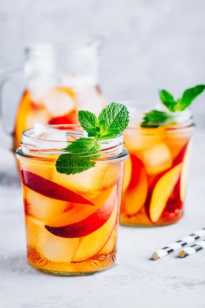 Homemade peach iced tea or lemonade with fresh mint and ice cubes in glass jar on gray stone background.