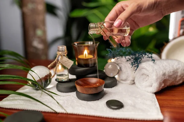 A girl pours oil from a jar of aromatic oils standing on stones for stone therapy and located on a terry towel next to which are transparent spheres, a twisted terry towel of white color and a sprig of lavender