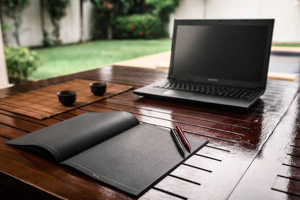 A wooden outdoor desk with a black notebook on it, two pens and clay cups for drinks