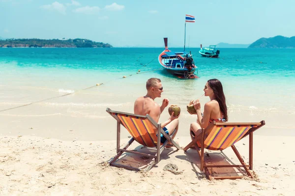 rear view of young couple with coconut cocktails relaxing in sun loungers on sandy beach.