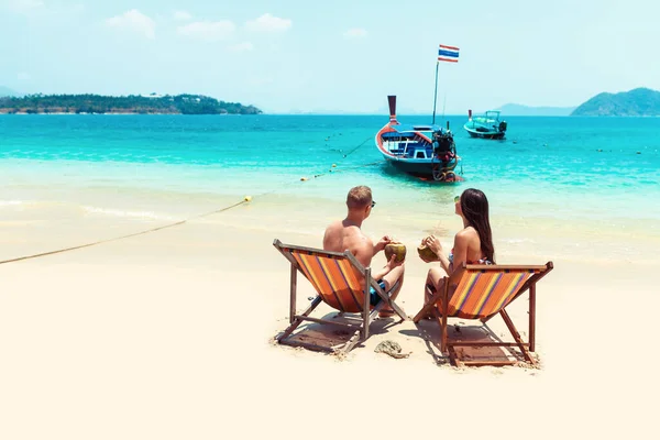 Rear view of young couple with coconut cocktails relaxing in sun loungers on sandy beach. — Stock Photo, Image