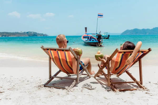 Rear view of young couple with coconut cocktails relaxing in sun loungers on sandy beach. Beautiful sea and boat — Stock Photo, Image