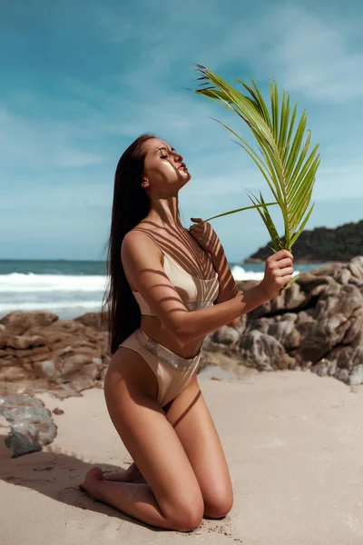 Foto de moda de mulher bonita com sardas e cabelo morena em um biquíni posando com verde uma folha de palma na praia — Fotografia de Stock