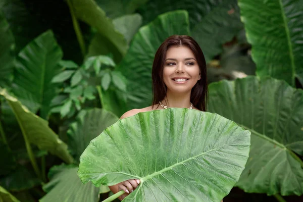 Une jeune brune avec une belle peau et un large sourire blanc neige pose avec une grande feuille verte. Soin biologique. — Photo