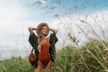 Omzunda örgülü uzun örgülü, yeşil ceketli ve sırtında kahverengi bir sırt çantasıyla tarlada poz veren seksi bir sarışın.