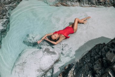 Çok güzel, ince, kırmızı bir mayo giymiş sarışın. Denizde, kayaların yanında yatıyor. Tropikal moda