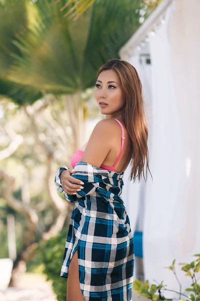 Retrato de rua de uma jovem morena asiática em um vestido xadrez de pé e posando no parque da cidade ao ar livre, com fundo de palmeiras borradas . — Fotografia de Stock