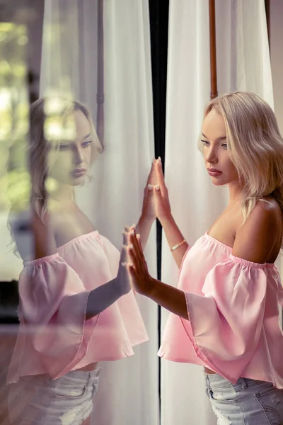 Delicate blonde in a trendy pink top touches the window with her reflection. The concept of style and beauty. — Stock Photo, Image