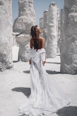 Bronzlaşmış, ince, makyajlı, siyah saçlı, beyaz kayaların yanında poz veren uzun beyaz elbiseli genç kadın. Arka plan,