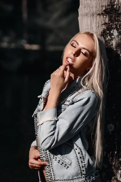 Foto de moda de la sexy modelo rubia en una chaqueta de mezclilla de pie cerca de la palmera. fondo de bosque tropical . —  Fotos de Stock