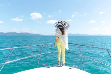 Arkadan bak. Yazlık takım elbiseli, şapkalı, yatın kenarında duran ve gezi sırasında güzel doğa manzarası seyreden güzel bir kadının açık hava fotoğrafı. Yaz seyahatinden zevk alan mutlu kadın. Arka plan