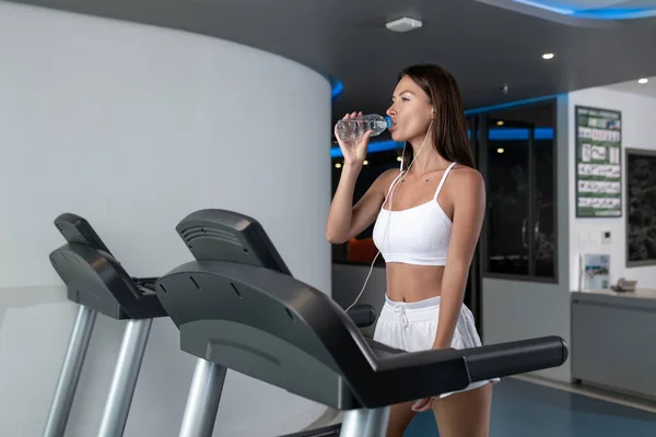 Athletic beautiful brunette in a white sports top and white short sports shorts, with headphones posing while running on a treadmill. Drinking water from a bottle. Sport. Fitness.