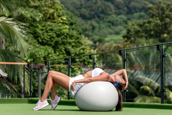 在室外 穿着蓝色耳机 躺在一个白色的运动球上 做运动女子的姿势 健康的天性 伸懒腰 — 图库照片