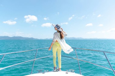 Arkadan bak. Yazlık takım elbiseli, şapkalı, yatın kenarında duran ve gezi sırasında güzel doğa manzarası seyreden güzel bir kadının açık hava fotoğrafı. Yaz seyahatinden zevk alan mutlu kadın. Arka plan