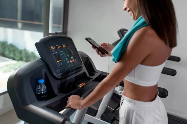 Athletic beautiful brunette in a white sports top and white short sports shorts, with headphones, with a blue towel on her neck posing on a treadmill, looking at the phone. Photo from the back.